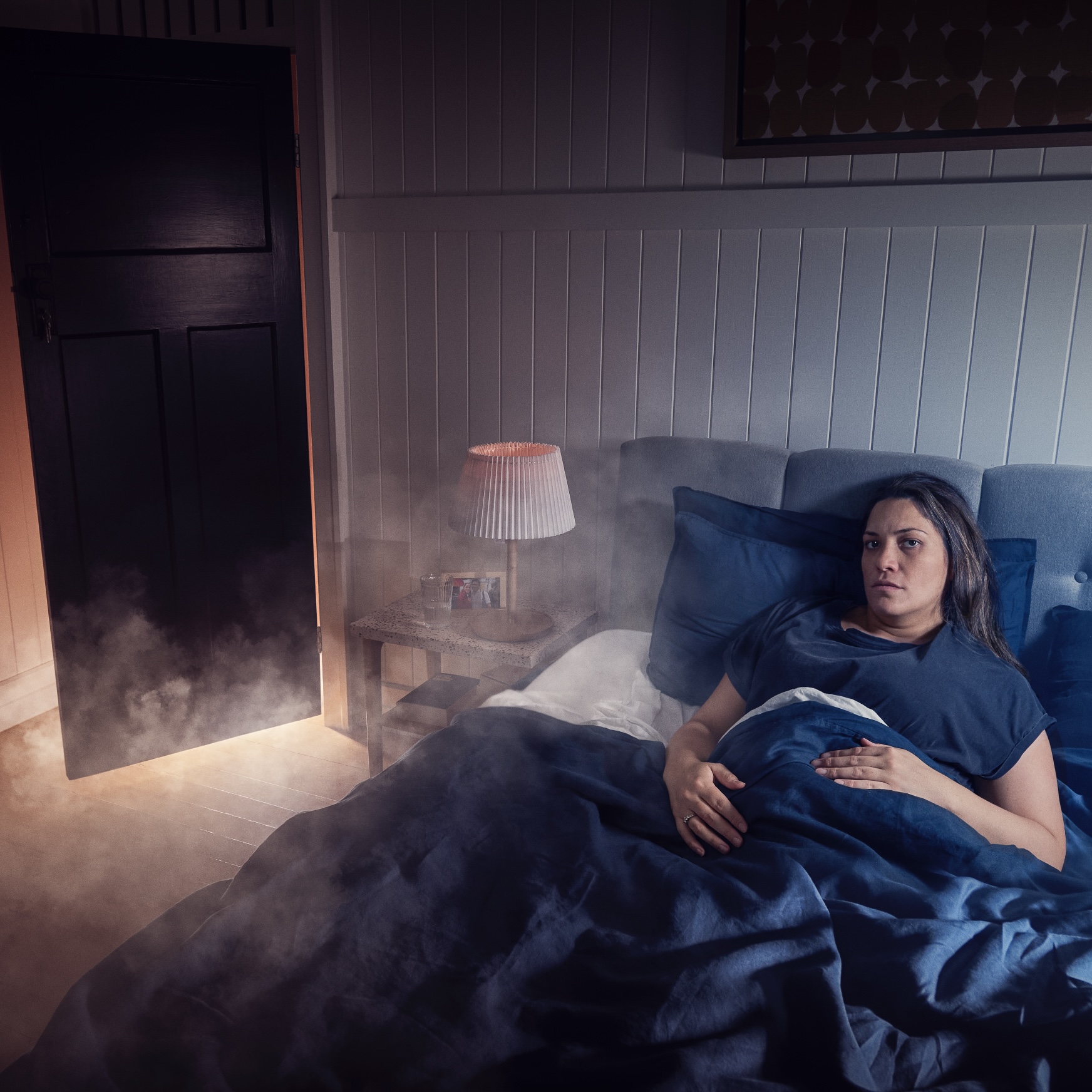 A woman lies in bed while smoke fills the bedroom.