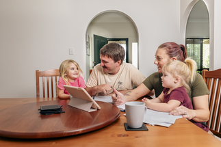 A young family with two children sit at the dining room table looking at a tablet.