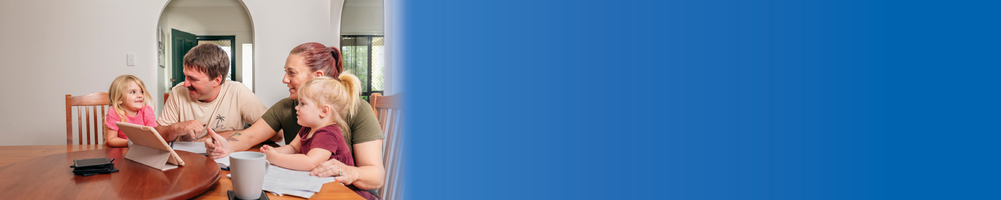 A young couple with two children sit at a dining room table looking at a tablet. 