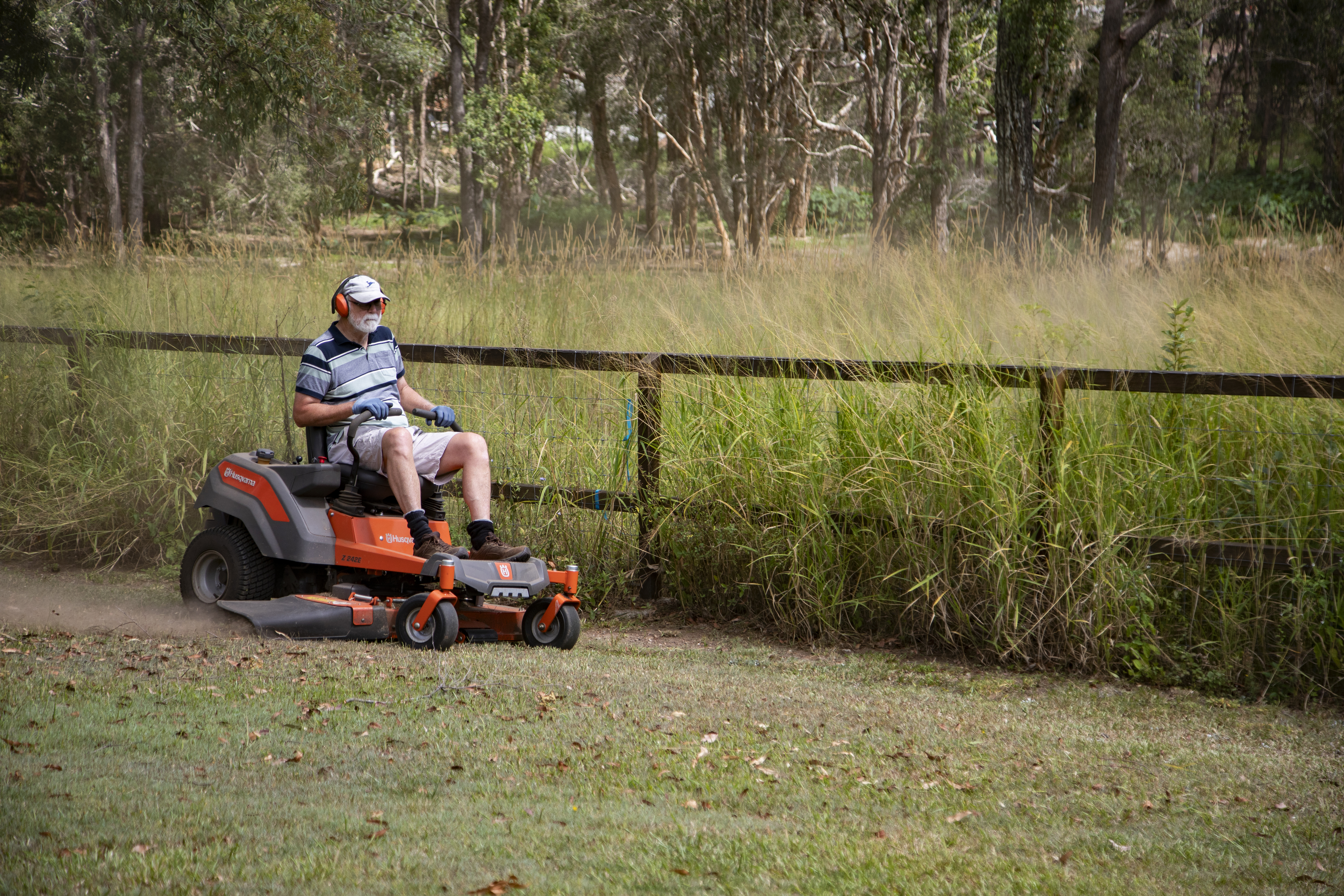 A man is on a ride on mower, mowing his yard. Long grass is in the background. 